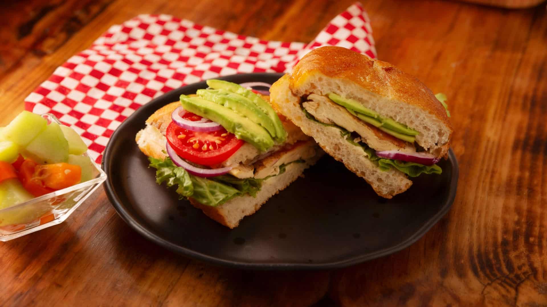 Fresh chicken sandwich with avocado, tomato, and lettuce on a rustic wooden table over checkered napkin, highlighting healthy ingredients and delicious flavor.